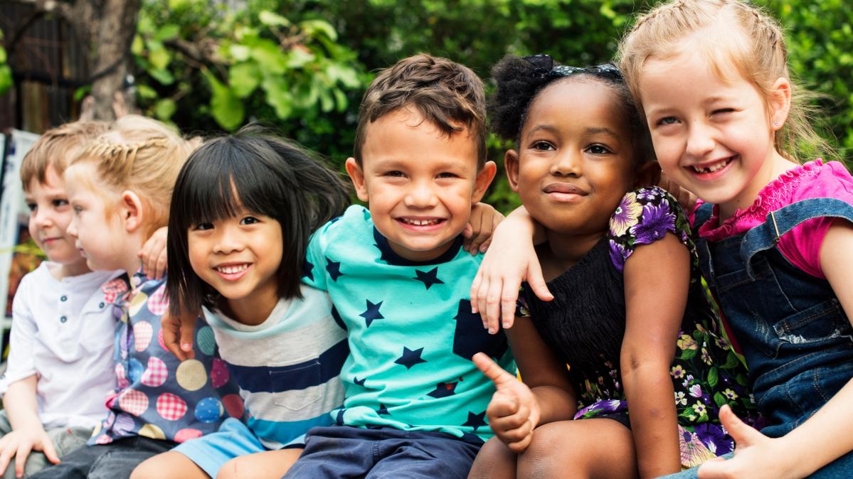 Six smiling, diverse pre-school children seated outdoors with arms draped around one another's shoulders.