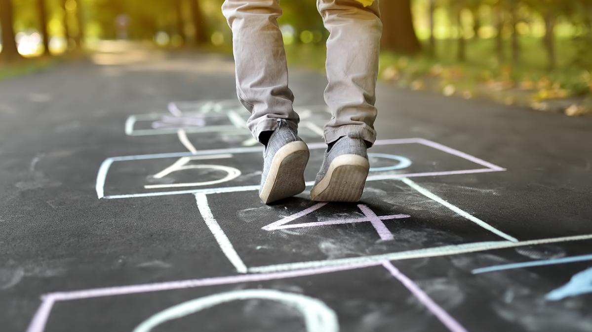 Looking at ground level at a white chalk hopscotch pattern on black paved path in a forest with only the lower legs and bottom of a yellow raincoat showing of a child stepping on a hopscotch square..