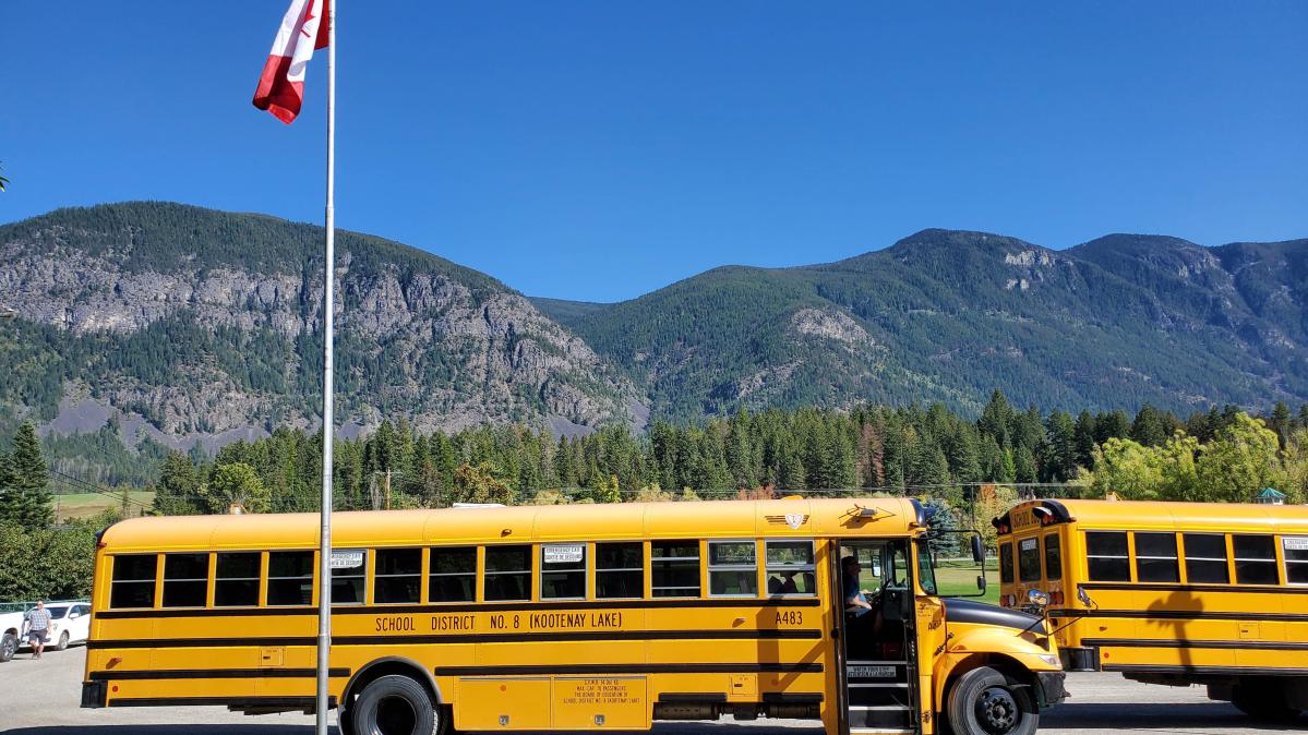 A yellow school bus in a parking lot with a Canadian flag in the foreground and mountains and cloudless blue sky in the background.