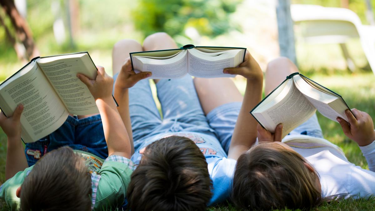 Three children lie side by side on their backs on the grass, each reading a book.