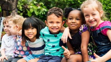 Six smiling, diverse pre-school children seated outdoors with arms draped around one another's shoulders.