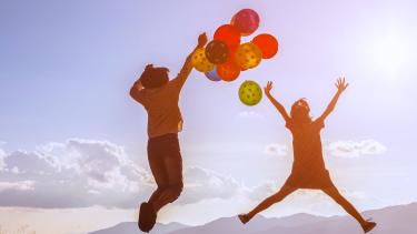 Two children leap into the air with coloured balloons against a background of mountains and sunshine.
