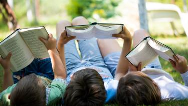 Three children lie side by side on their backs on the grass, each reading a book.
