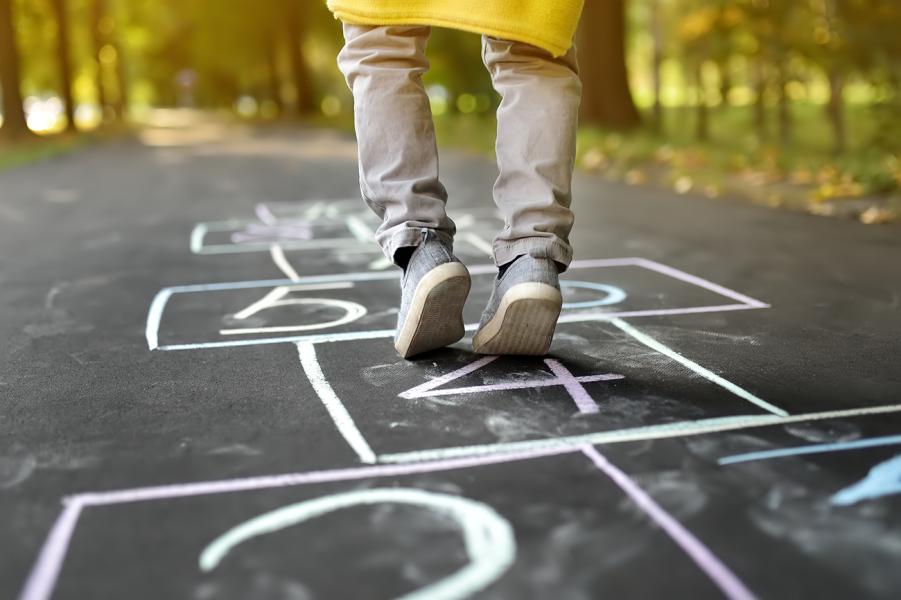 Looking at ground level at a white chalk hopscotch pattern on black paved path in a forest with only the lower legs and bottom of a yellow raincoat showing of a child stepping on a hopscotch square..